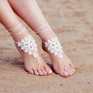May include: White crochet foot jewelry with delicate lace patterns. The jewelry is worn on both feet and is shown on a sandy beach.