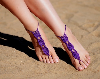 Op de afbeelding: Paarse gehaakte voetsieraden met ingewikkelde ontwerpen. De sieraden worden op beide voeten gedragen en worden getoond op een zanderig strand.