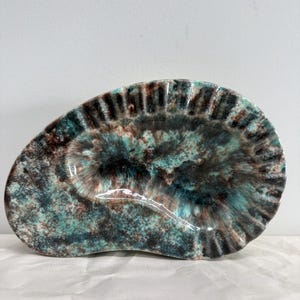 Könnte beinhalten: Eine ovale Keramikschale mit strukturierter Oberfläche. Die Schale hat eine Mischung aus teal, braunen und schwarzen Farben, wodurch ein Marmoreffekt entsteht. Der Rand hat ein geriffeltes Design. Dieses dekorative Stück eignet sich zum Servieren oder Ausstellen.