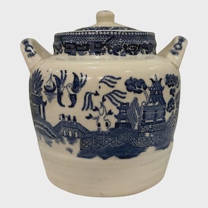 Puede incluir: Un tarro de cerámica blanco con tapa, con un diseño azul y blanco de patrón Willow. El tarro tiene dos asas y un pequeño pomo en la tapa. El patrón representa un paisaje con puentes, pagodas y figuras.