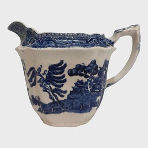 Puede incluir: Una jarra de cerámica blanca y azul con un diseño detallado de patrón Willow. La jarra tiene un asa curva y un pico, con un borde decorativo alrededor del borde. El patrón azul representa una escena de paisaje.