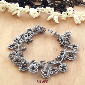 Puede incluir: Un brazalete de crochet plateado con cuentas de color gris oscuro. El brazalete está hecho de delicadas hojas de crochet y tiene un diseño floral.