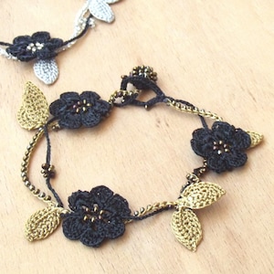 Puede incluir: Un brazalete de crochet negro con hojas de crochet doradas y cuentas negras. El brazalete está hecho de un delicado y complejo trabajo de crochet.