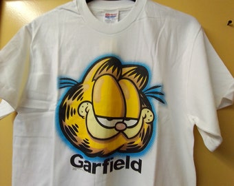 May include: White t-shirt with a cartoon image of Garfield the cat. The image is in blue, yellow, and black. The text "Garfield" is printed below the image.