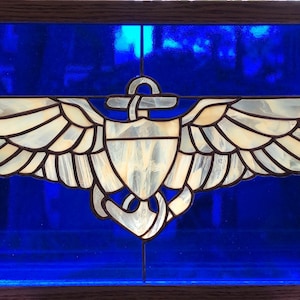 May include: A stained glass window depicting a naval aviator's wings with an anchor in the center. The wings are white with gold trim and the anchor is silver. The background is a deep blue.
