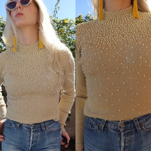 以下が含まれることがあります： モックネックとパールがあしらわれたベージュのリブニットセーター。長袖で、柔らかく快適な素材で作られています。