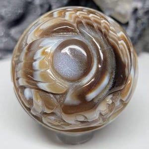 May include: A polished spherical stone carving featuring intricate, swirling patterns in shades of brown, tan, and white. The surface displays a smooth, glossy finish with a prominent circular focal point at the center, showcasing natural banding.