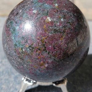 May include: A polished spherical stone featuring a mottled texture with deep magenta, dark green, and metallic gold inclusions. The sphere rests on a small, three-legged silver-toned metal stand, showcasing a smooth, reflective surface throughout.