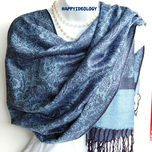 May include: A blue paisley patterned scarf with a fringed edge. The scarf is draped over a mannequin, showcasing its intricate design and color variations. The scarf has a light blue and dark blue color scheme.