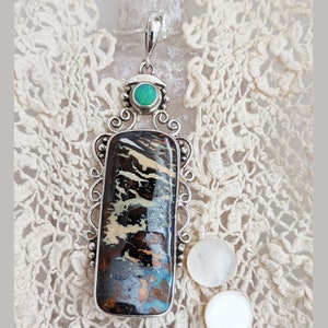 May include: A silver pendant with a large, rectangular, black, white, and blue opal stone. The pendant has a smaller, round, green stone at the top. The pendant is on a white lace background.