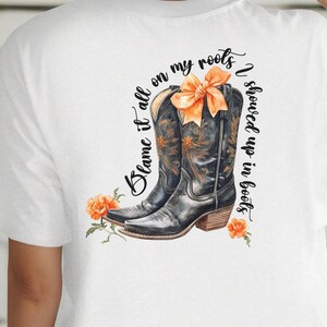 May include: White t-shirt with a graphic of brown cowboy boots with orange flowers and the text "Blame it all on my roots I showed up in boots."