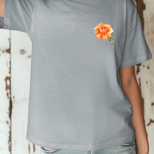 Op de afbeelding: Een lichtgrijs T-shirt met een kleine, oranje geborduurde roos op de linkerborst.