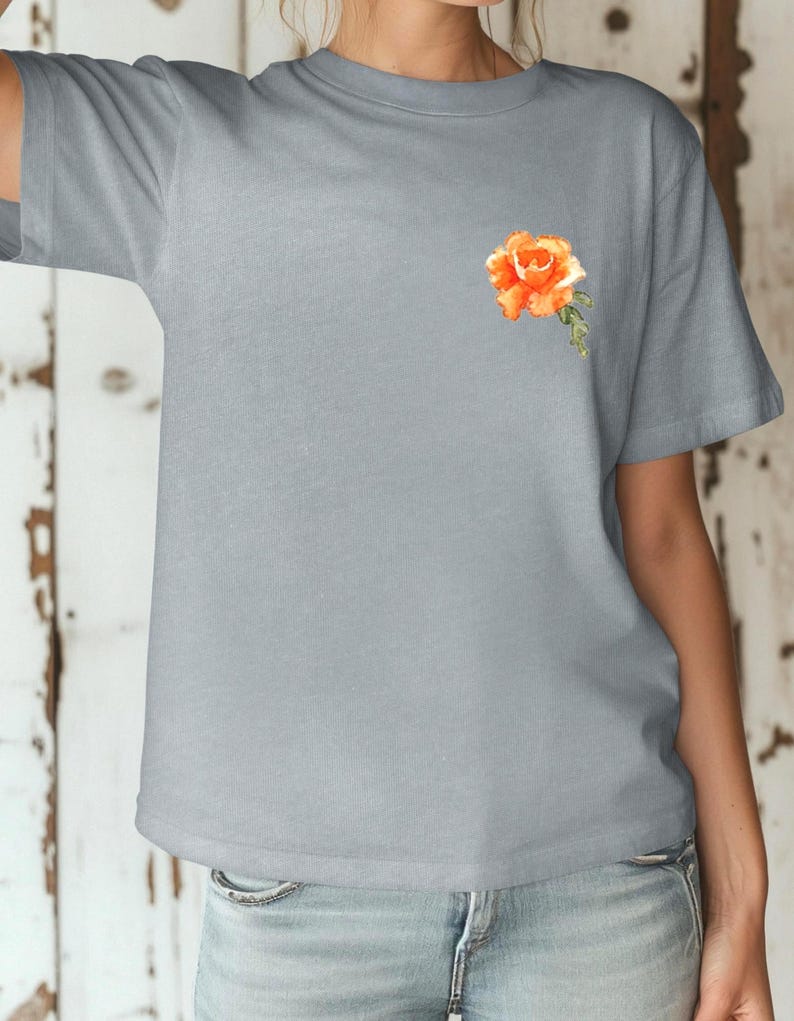 Op de afbeelding: Een lichtgrijs T-shirt met een kleine, oranje geborduurde roos op de linkerborst.