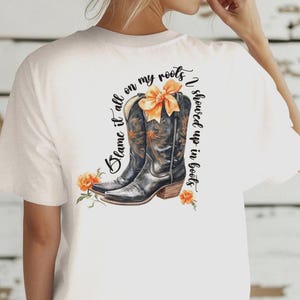 Op de afbeelding: Wit T-shirt met een zwarte en bruine cowboylaars afbeelding en de tekst "Blame it all on my roots I showed up in boots".