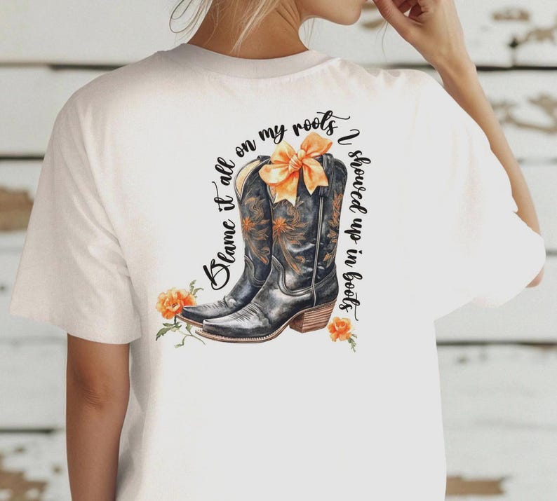 Op de afbeelding: Wit T-shirt met een zwarte en bruine cowboylaars afbeelding en de tekst "Blame it all on my roots I showed up in boots".