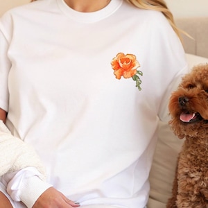 Op de afbeelding: Wit T-shirt met een oranje geborduurde roos op de linker borst.