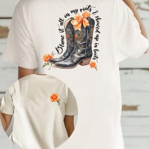 Op de afbeelding: Wit T-shirt met een afbeelding van een paar bruine cowboylaarzen met oranje bloemen en de tekst "Blame it all on my roots I showed up in boots".