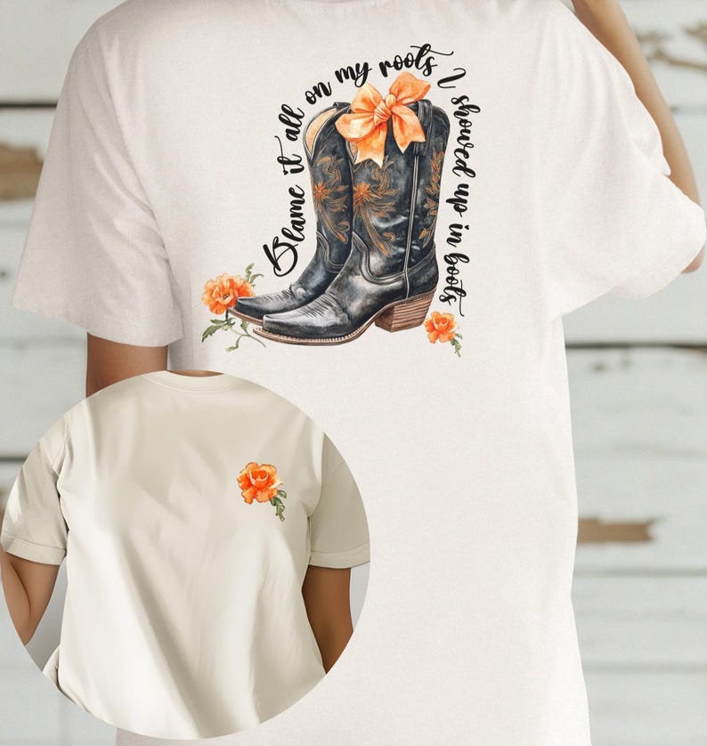 Op de afbeelding: Wit T-shirt met een afbeelding van een paar bruine cowboylaarzen met oranje bloemen en de tekst "Blame it all on my roots I showed up in boots".