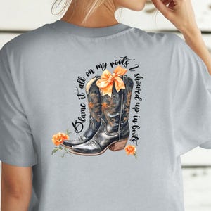 Op de afbeelding: Een lichtgrijs T-shirt met een afbeelding van twee zwarte cowboylaarzen met een oranje strik. De tekst "Blame it all on my roots I showed up in boots" is op het T-shirt gedrukt.