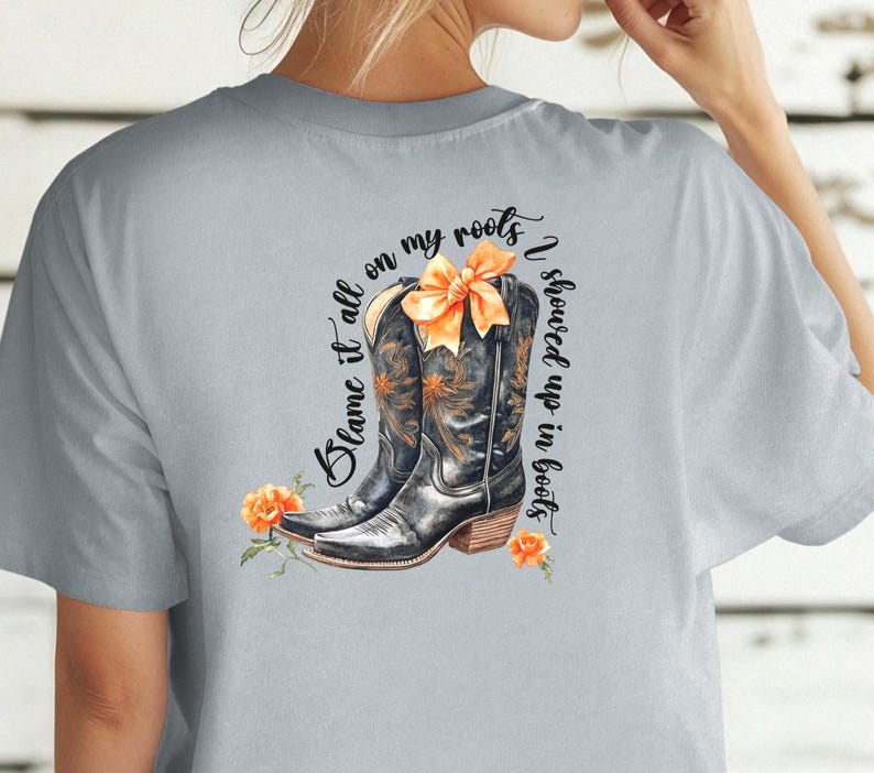 Op de afbeelding: Een lichtgrijs T-shirt met een afbeelding van twee zwarte cowboylaarzen met een oranje strik. De tekst "Blame it all on my roots I showed up in boots" is op het T-shirt gedrukt.