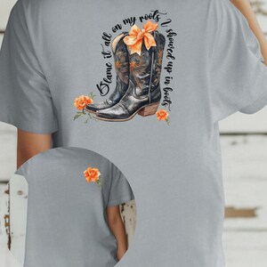 Op de afbeelding: Een grijs T-shirt met de tekst "Blame it all on my roots I showed up in boots" en een paar zwarte cowboylaarzen met een oranje strik. De laarzen zijn omringd door oranje bloemen.