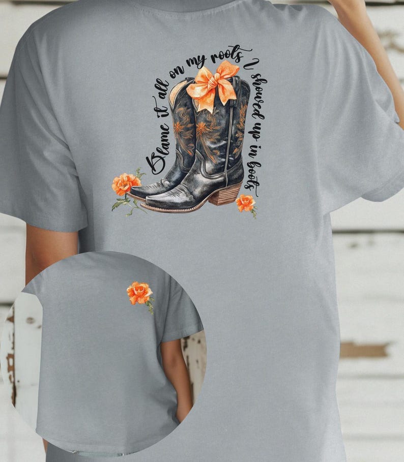 Op de afbeelding: Een grijs T-shirt met de tekst "Blame it all on my roots I showed up in boots" en een paar zwarte cowboylaarzen met een oranje strik. De laarzen zijn omringd door oranje bloemen.