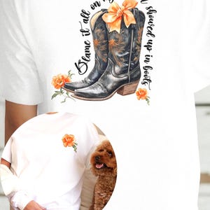 Op de afbeelding: Wit T-shirt met een paar zwarte cowboylaarzen met een oranje strik. De tekst "Blame it all on my roots I showed up in boots" staat op het T-shirt gedrukt.