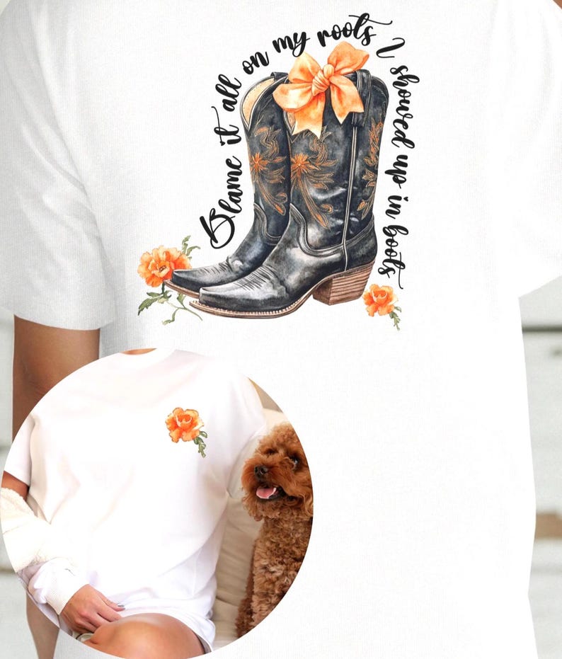 Op de afbeelding: Wit T-shirt met een paar zwarte cowboylaarzen met een oranje strik. De tekst "Blame it all on my roots I showed up in boots" staat op het T-shirt gedrukt.