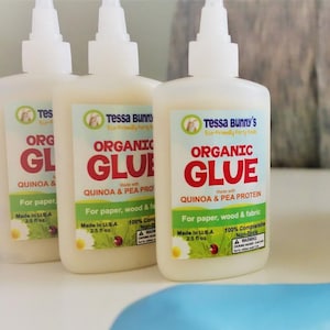 Può includere: Tre bottiglie bianche di colla organica Tessa Bunny's. Le etichette presentano il testo "Organic Glue" in rosso, con "Quinoa & Pea Protein" e "For paper, wood & fabric" sotto. Ogni bottiglia contiene 74 ml.