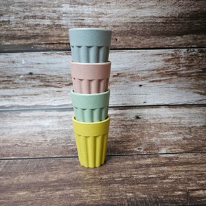 May include: Four stackable cups made of bamboo in yellow, green, pink, and blue colors. The cups are stacked on a wooden surface.