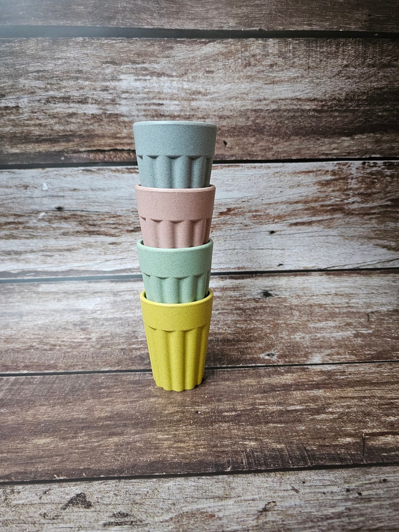 May include: Four stackable cups made of bamboo in yellow, green, pink, and blue colors. The cups are stacked on a wooden surface.