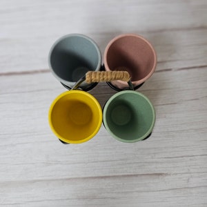 May include: Four colorful cups in a metal holder with a rope handle. The cups are light blue, pink, yellow, and green.