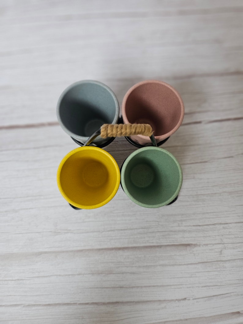 May include: Four colorful cups in a metal holder with a rope handle. The cups are light blue, pink, yellow, and green.