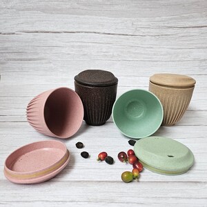 May include: An assortment of reusable coffee cups and lids in various colors, including pink, brown, and green. The cups have a textured, ribbed design. Coffee beans and berries are scattered around the cups.