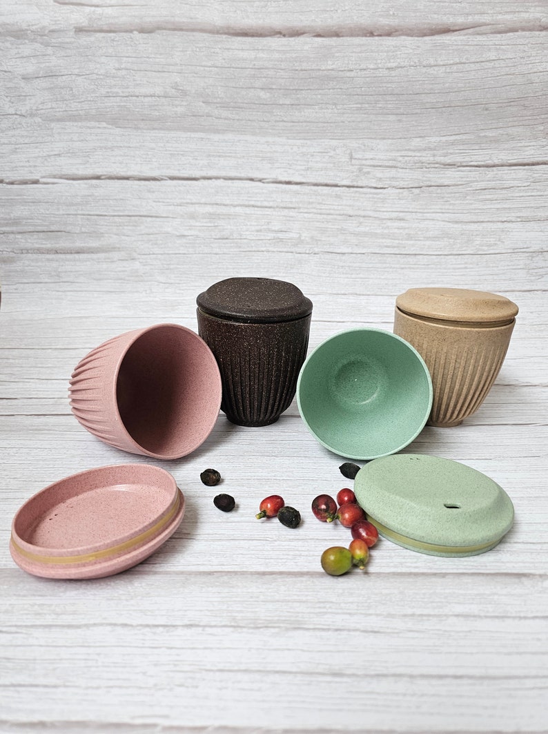 May include: An assortment of reusable coffee cups and lids in various colors, including pink, brown, and green. The cups have a textured, ribbed design. Coffee beans and berries are scattered around the cups.