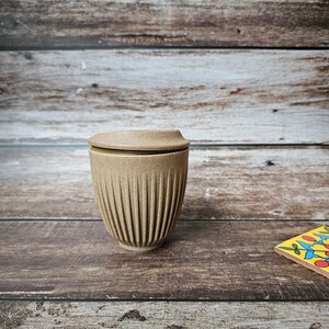 May include: A tan, ribbed ceramic travel mug with a matching lid. The mug is cylindrical with vertical grooves and sits on a wooden surface. The lid is flat and fits snugly on top. The background is a wooden wall.