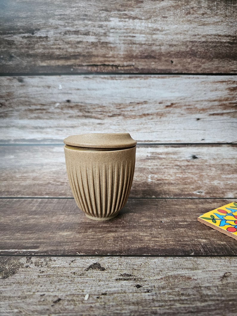 May include: A tan, ribbed ceramic travel mug with a matching lid. The mug is cylindrical with vertical grooves and sits on a wooden surface. The lid is flat and fits snugly on top. The background is a wooden wall.