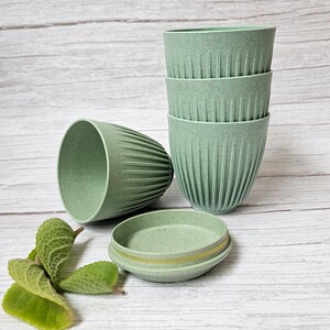 May include: A set of four light green, ribbed cups stacked together, with a fifth cup and a small, round container with a lid nearby. The cups have a textured appearance. Green leaves are in the foreground. The background is a light wood grain.