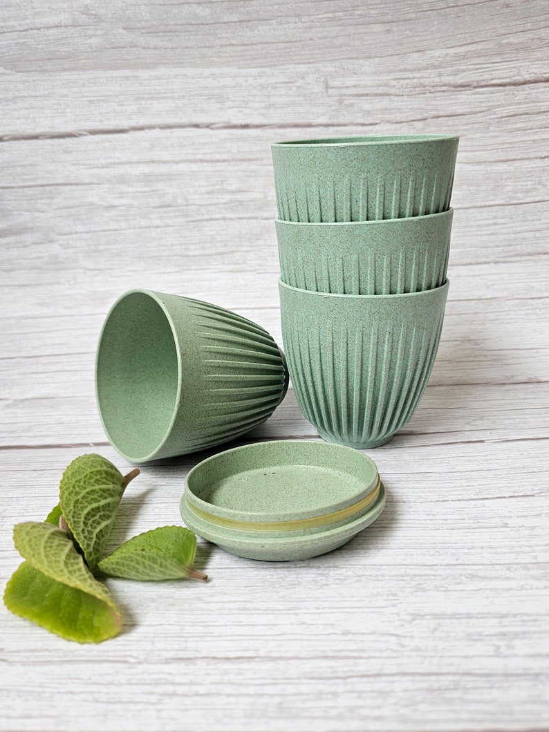 May include: A set of four light green, ribbed cups stacked together, with a fifth cup and a small, round container with a lid nearby. The cups have a textured appearance. Green leaves are in the foreground. The background is a light wood grain.