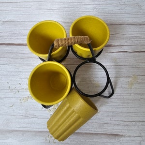 May include: Set of four yellow ceramic mugs with a black metal stand and a rope handle. The mugs are arranged in a circle with the stand in the center. The stand has a ring on the top for hanging the mugs.
