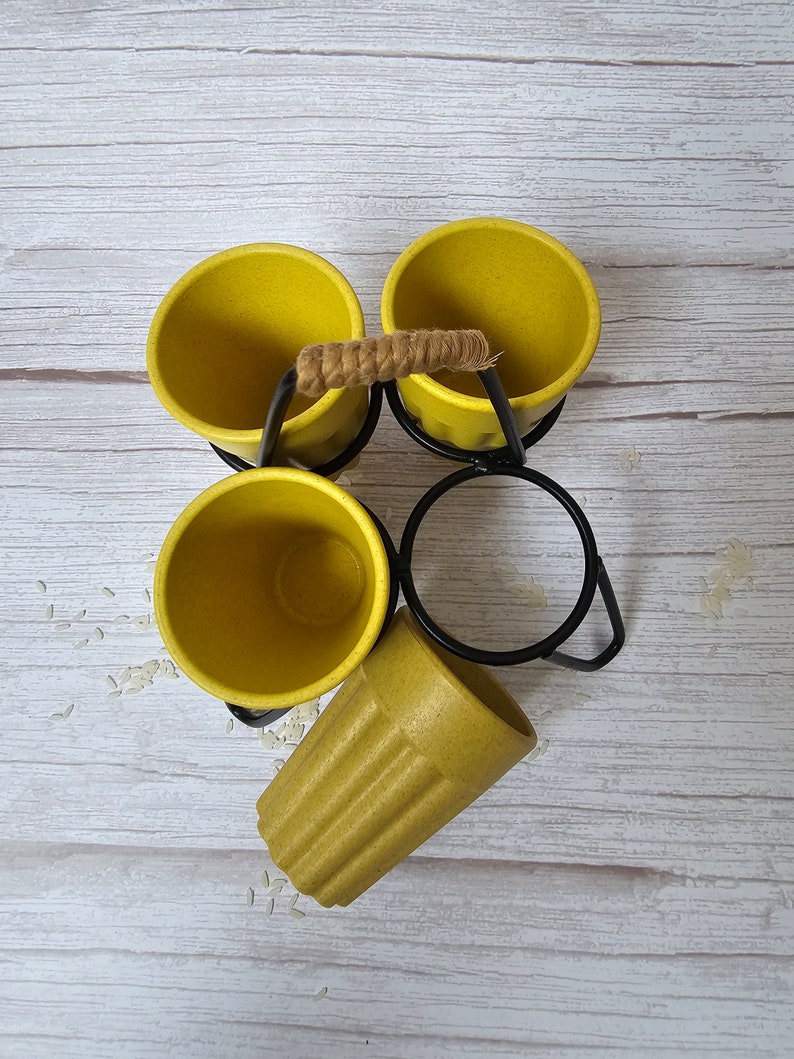 May include: Set of four yellow ceramic mugs with a black metal stand and a rope handle. The mugs are arranged in a circle with the stand in the center. The stand has a ring on the top for hanging the mugs.