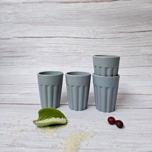 May include: Four gray ceramic cups with a ribbed design, stacked in a pyramid shape. The cups are on a white wooden surface with a green leaf and white rice scattered around them.