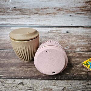 May include: Two reusable coffee cups with lids. One cup is beige with vertical ridges, and the other is pink. Both cups are made of a speckled material. The cups are on a wooden surface.