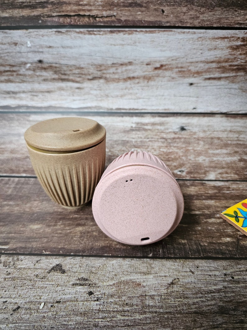 May include: Two reusable coffee cups with lids. One cup is beige with vertical ridges, and the other is pink. Both cups are made of a speckled material. The cups are on a wooden surface.