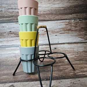 May include: A set of four stackable cups with a black metal stand. The cups are made of a light brown material and are in the colors pink, green, yellow, and gray.