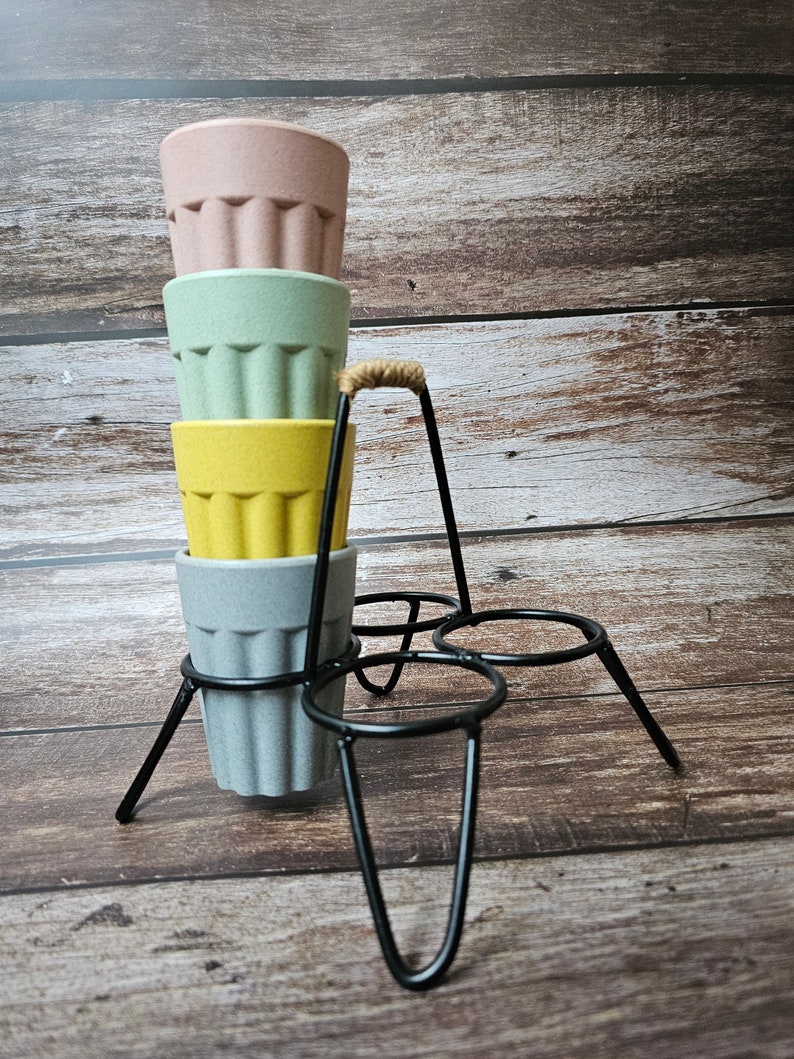 May include: A set of four stackable cups with a black metal stand. The cups are made of a light brown material and are in the colors pink, green, yellow, and gray.