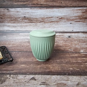 May include: A light green ceramic container with a matching lid. The container has vertical ribbed detailing and a cylindrical shape. The lid is flat and round. The container is on a wooden surface.