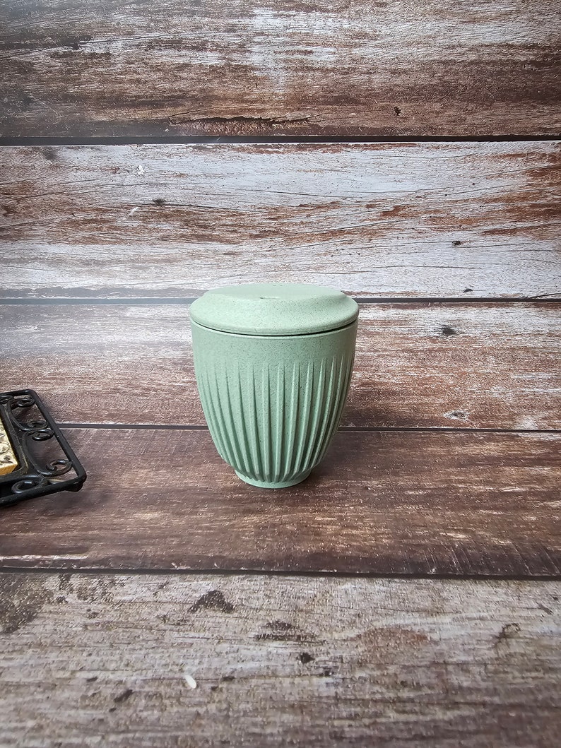May include: A light green ceramic container with a matching lid. The container has vertical ribbed detailing and a cylindrical shape. The lid is flat and round. The container is on a wooden surface.