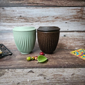 May include: Two reusable coffee cups with lids. One is light green, the other is brown. Both cups have a ribbed design. The cups are on a wooden surface with small decorative items.