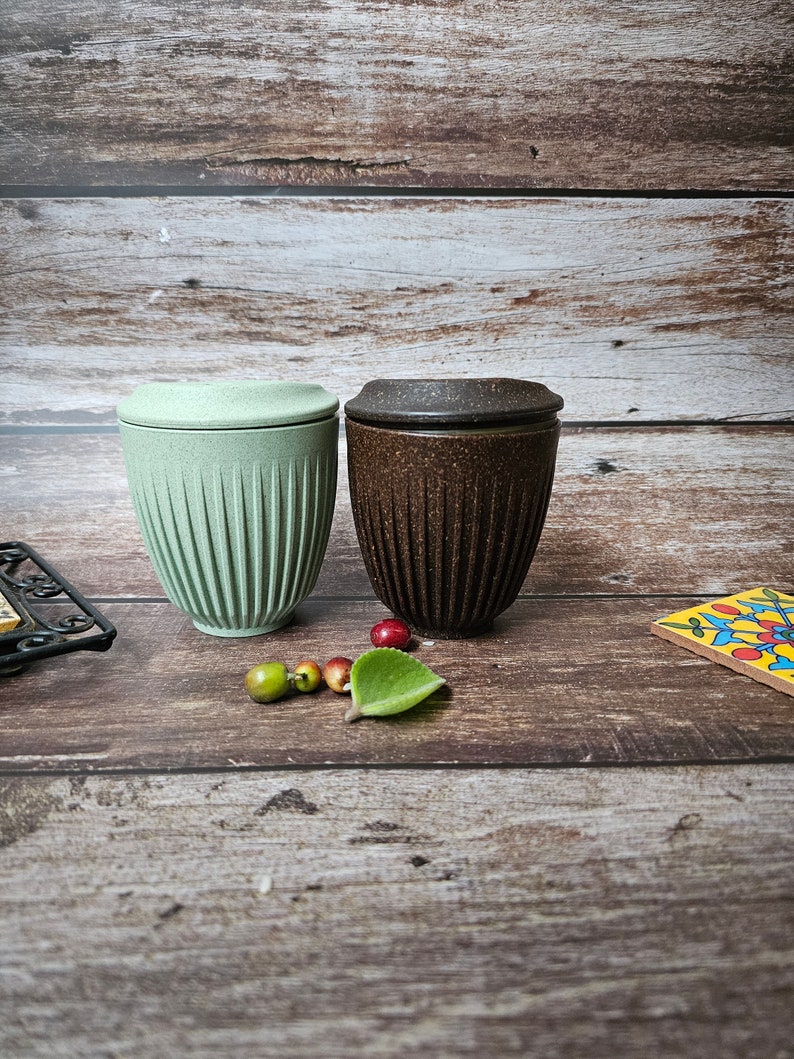 May include: Two reusable coffee cups with lids. One is light green, the other is brown. Both cups have a ribbed design. The cups are on a wooden surface with small decorative items.